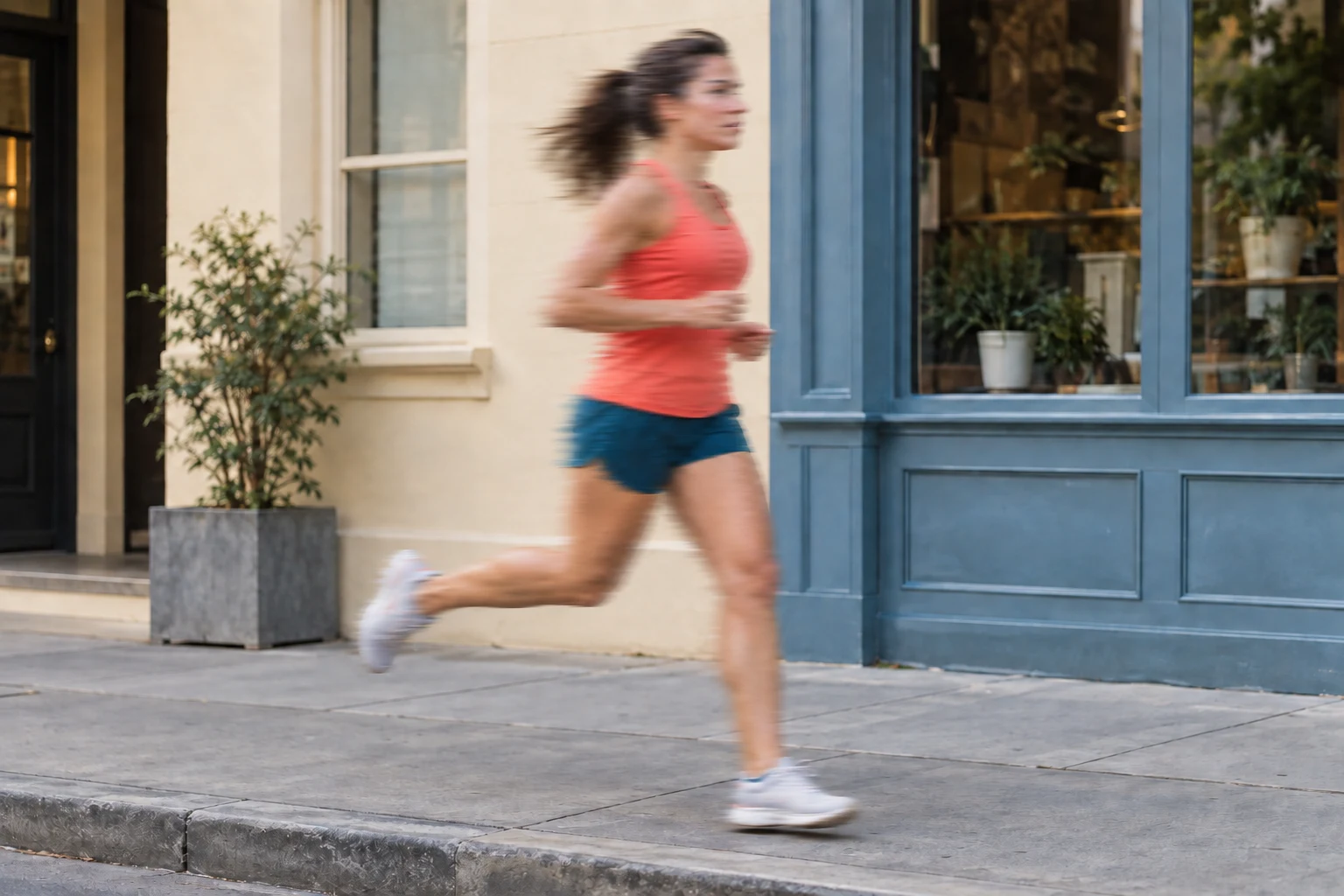 Full-body view of a woman in a coral athletic top and dark teal shorts running along a city sidewalk — Blurred